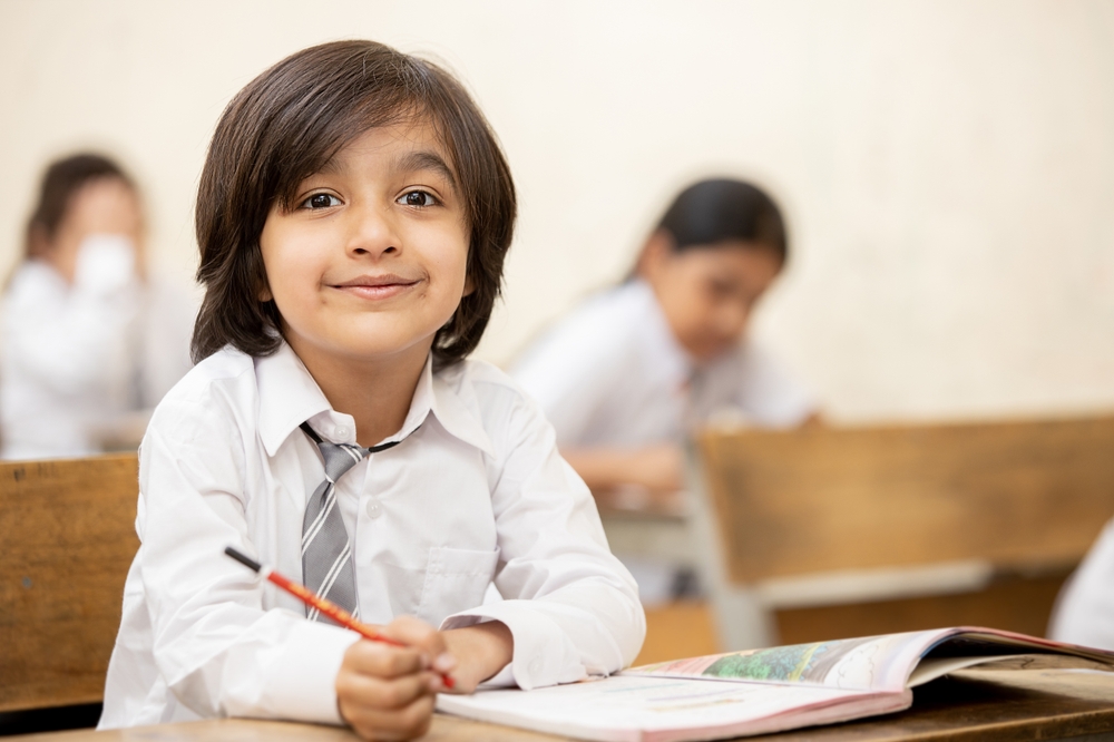 Portrait,Of,Happy,Indian,School,Sitting,At,Desk,In,Classroom,